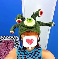 a woman with red hair wearing a green knitted frog hat and holding a heart