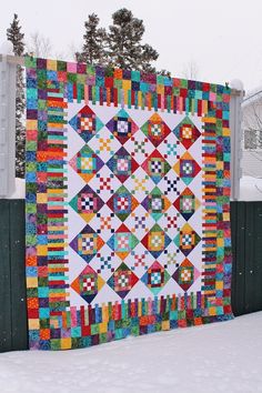 a colorful quilt is hanging on a fence in the snow with pine trees behind it