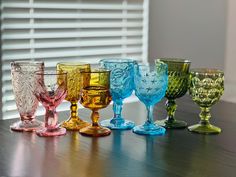 several different colored glass goblets lined up on a wooden table in front of a window