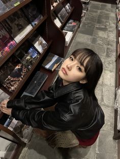 a woman in a black leather jacket is looking up at the camera and she's leaning on a bookshelf