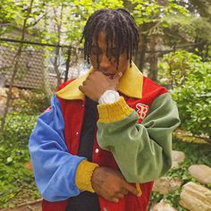 a man with dreadlocks standing in front of some rocks and trees, holding his hand to his face