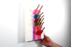 a hand is holding an art piece with popsicles on it and pink, green, blue, and red sticks sticking out of it