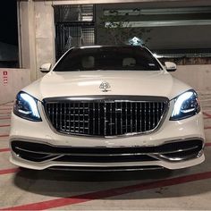 the front end of a white car parked in a parking lot with its lights on