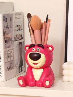 a pink bear with makeup brushes in it's mouth sitting on a shelf next to a book