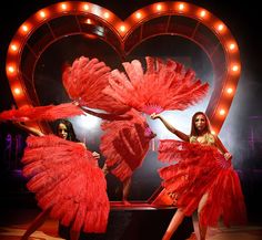 two women dressed in red are dancing on stage with lights behind them and a heart shaped sign