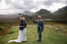 a man and woman standing on top of a lush green hillside holding up an umbrella