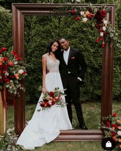 a bride and groom standing in front of a wooden frame with red flowers on it