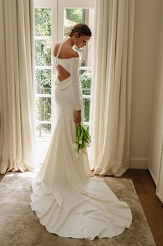 a woman standing in front of a window wearing a white wedding dress and holding flowers