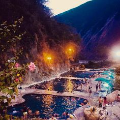 many people are swimming in the pool at night with lights shining on them and mountains in the background