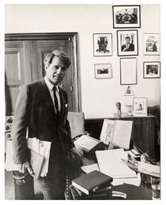 black and white photograph of man in suit standing next to desk with papers on it