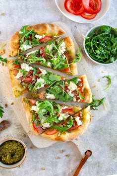 two pizzas with different toppings sit on a cutting board next to bowls of pesto and tomatoes