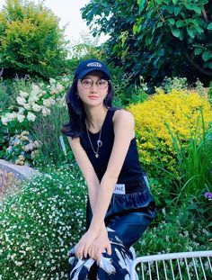 a woman sitting on top of a metal bench in front of flowers and bushes with her hand under her chin