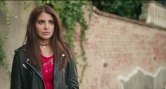 a woman in a red shirt and black jacket standing next to a brick wall with ivy growing on it