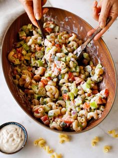 two hands are scooping macaroni salad from a wooden bowl and another hand is holding a spoon