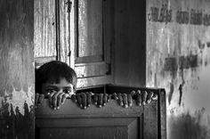 a young boy peeking out from behind a wooden door with his hands on top of the door