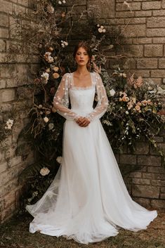 a woman standing in front of a brick wall wearing a wedding dress with sheer sleeves