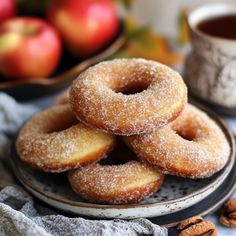 some sugared doughnuts are on a plate with coffee and apples in the background