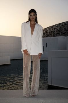 a woman standing in front of a body of water wearing a white jacket and pants