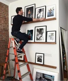 a man sitting on top of a ladder next to a wall filled with framed pictures