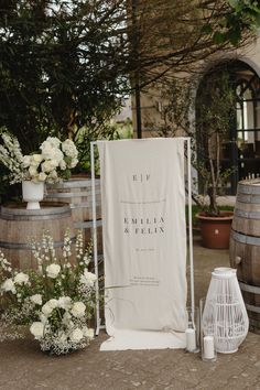 a white sign sitting on top of a wooden barrel next to flowers and vases