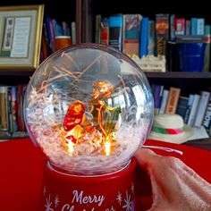 a hand holding a snow globe with christmas decorations in it on top of a red table