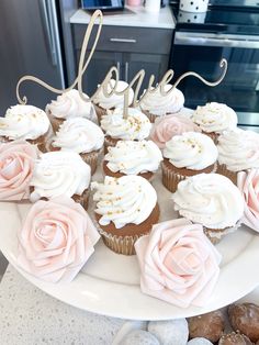 cupcakes with white frosting and pink roses are arranged on a cake plate