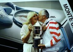 an older couple kissing each other while holding a trophy in front of a poster with tennis racquets on it