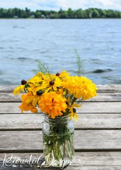 a mason jar filled with yellow flowers sitting on top of a wooden table next to the water