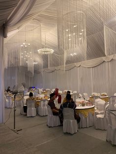 several people sitting at tables in a room with white drapes on the walls and chandeliers hanging from the ceiling