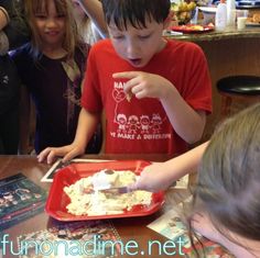 two children eating food at a table with other kids in the background and one child holding a fork