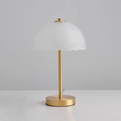 a white lamp with a gold base on a table top in front of a gray wall