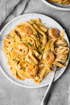 two white plates filled with pasta and shrimp on top of a gray tablecloth next to silver spoons