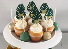 cupcakes with white frosting and green decorations on a cake platter, surrounded by greenery