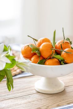 a white bowl filled with oranges sitting on top of a wooden table next to flowers