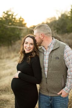 a pregnant woman and man standing next to each other in an open field at sunset