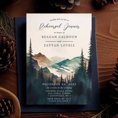 an image of a wedding card with mountains and pine cones on the table next to it