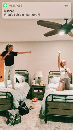 two people standing on top of twin beds in a room with ceiling fan and other items