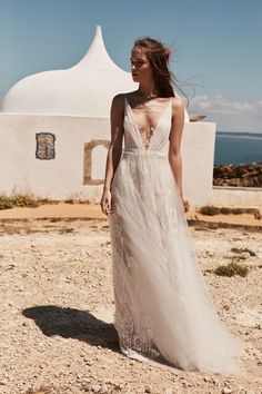 a woman in a long white dress standing on the beach with her back to the camera