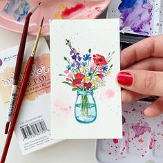 a hand holding a card with watercolor paints and flowers in a vase on it