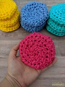 four crocheted round scrubcloths in different colors on a wooden surface with a hand holding one