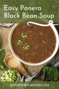 a bowl of black bean soup on top of a wooden table with green leaves and spoons