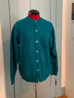 "This classic pocket cardigan by McKenzy is made of green-teal virgin wool. It has two pockets on the front and closes with 7 pearlescent plastic buttons. It has a spare button sewn inside. It measures approximately 45\" unstretched at the chest. The center back length is approximately 27\", the sleeves are approximately 23\" and the neck to shoulder is approximately 7\". It is in good condition. There is some discoloration on the underside of the sleeves. I am not sure if it is a stain. (It almost looks like dye settling from being washed.)" Classic Green Wool Cardigan, Green Button-up Cardigan With Pockets, Green Crew Neck Sweater With Pockets, Green Long Sleeve Sweater With Pockets, Pocket Cardigan, Sewing A Button, Wool Sweater, Wool Sweaters, Sweater Outfits