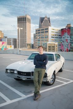 a man standing next to a white car in a parking lot with tall buildings behind him