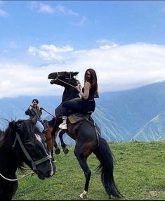 two women are riding horses on a grassy hill with mountains in the backgroud