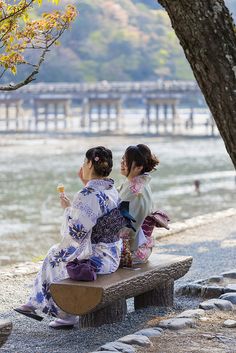 two women in kimonos sitting on a bench by the water eating ice cream