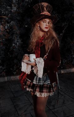 a woman in a skirt and top hat holding a coffee cup while standing next to a bush