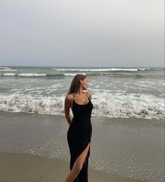 a woman in a black dress standing on the beach next to the ocean and waves