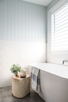 a white bath tub sitting under a window next to a wooden table with a potted plant on it