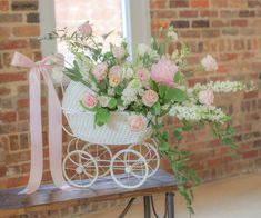 a white baby carriage with pink flowers and greenery on a wooden table in front of a brick wall