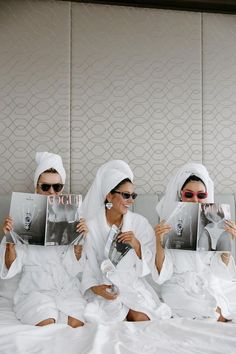 three women in white robes are sitting on a bed and one is reading a magazine